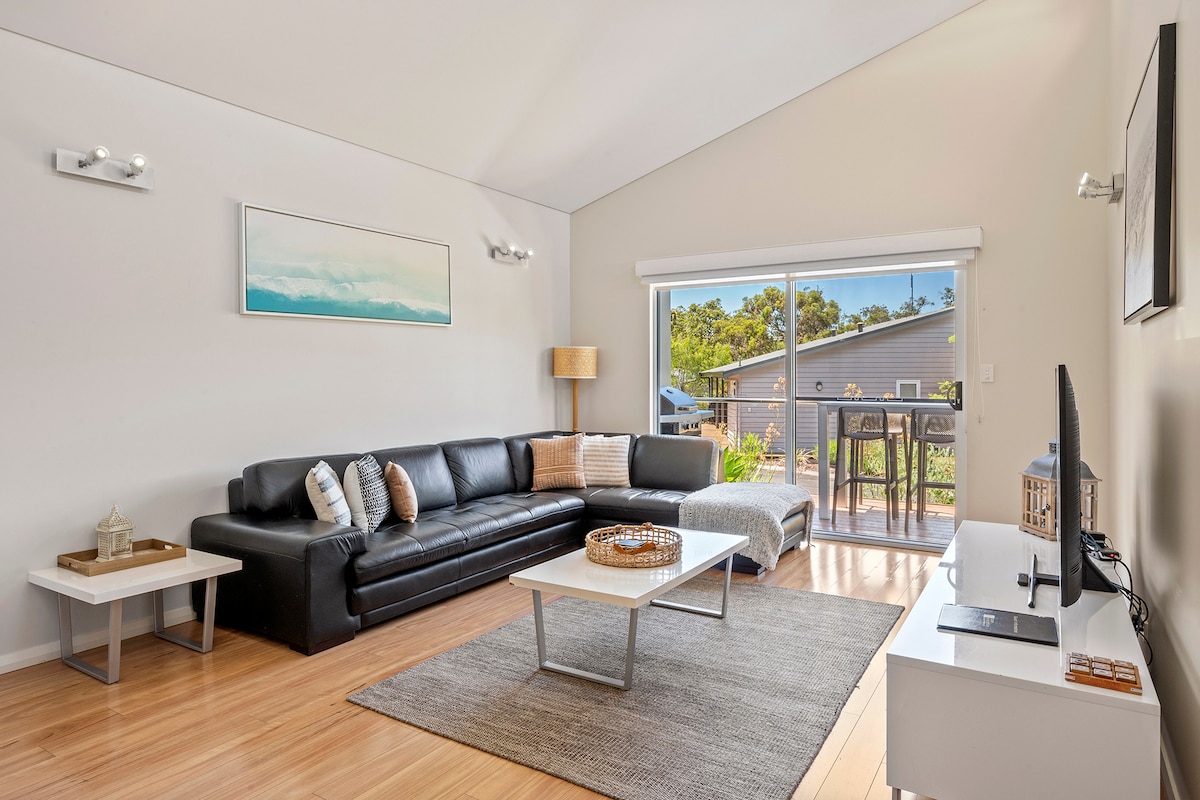 The open living area features a large black sectional sofa and a light-colored rug covering the wooden floor. Natural light floods through sliding glass doors, leading to a small deck. A coffee table and a sleek media console complete the serene space.