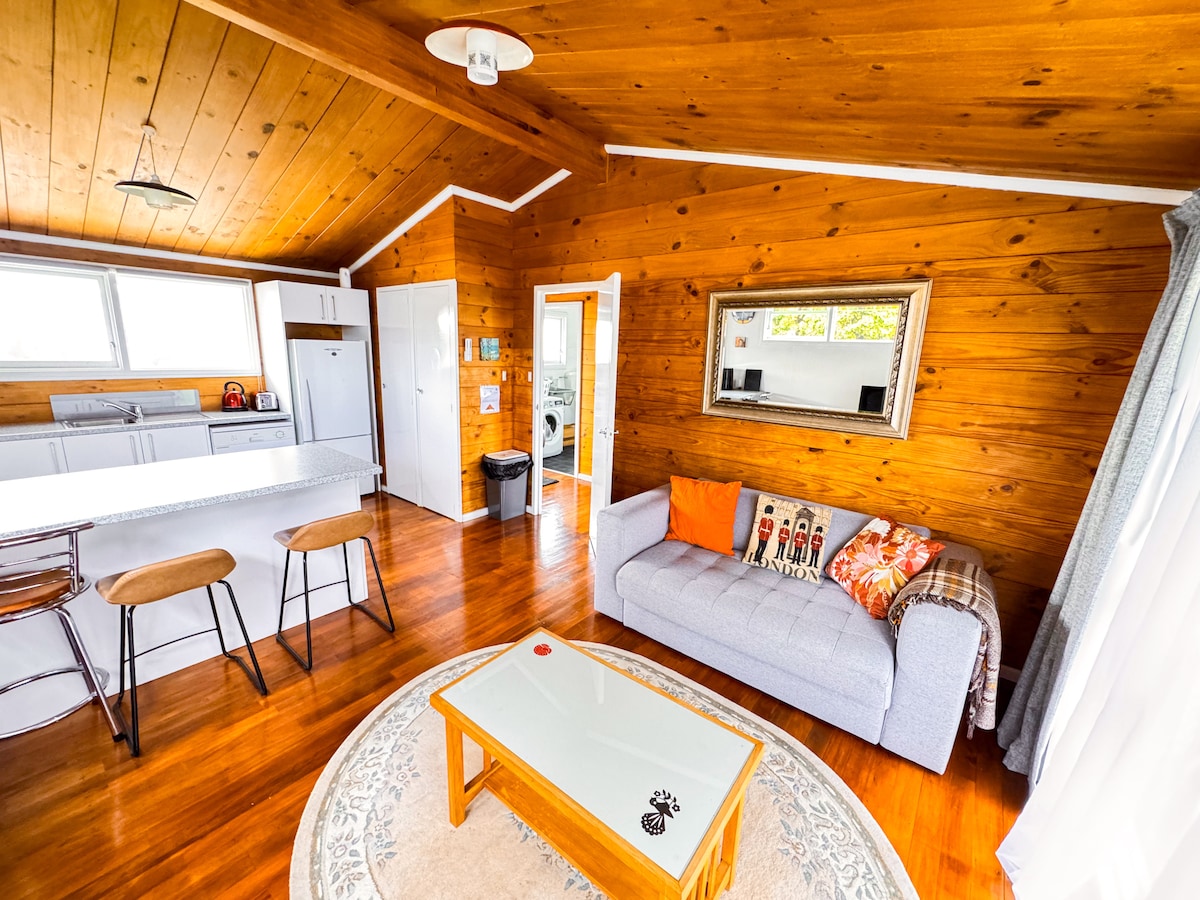 A bright living area features wooden paneling and polished floors. A comfortable sofa is adorned with decorative cushions, while a round coffee table sits nearby. A dining counter with two stools is positioned near the kitchen, which is partially visible through an open space.