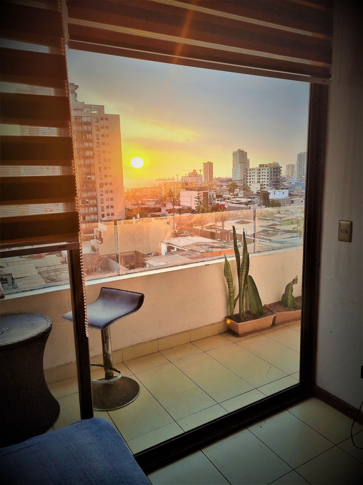 A balcony offers a view of the sunset over the city skyline, framed by large glass doors. A modern bar stool and a small round table are positioned on the balcony, complemented by potted plants beside them. Soft light enhances the warm tones of the evening sky.