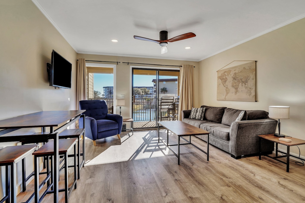 An open-concept living area features a comfortable sofa and a cozy blue armchair, with large sliding glass doors providing natural light and views of the balcony. A wall-mounted TV and a bar height table with stools complement the space, which is enhanced by a ceiling fan.