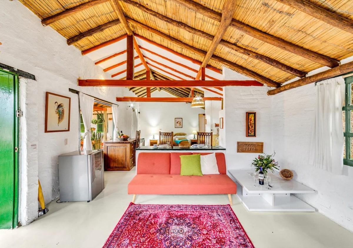 A spacious living area features a high bamboo ceiling with wooden beams and a vibrant area rug. A comfortable couch with green and white cushions complements the open layout, which includes a kitchenette and dining area in the background. Natural light filters through nearby windows.
