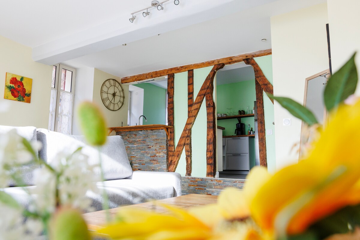 A spacious living area is captured with natural light illuminating the soft gray sofas. A rustic wooden feature frames the entrance to the kitchen, which is visible in the background. Bright, vibrant flowers are arranged on a wooden table, adding a touch of color to the scene.