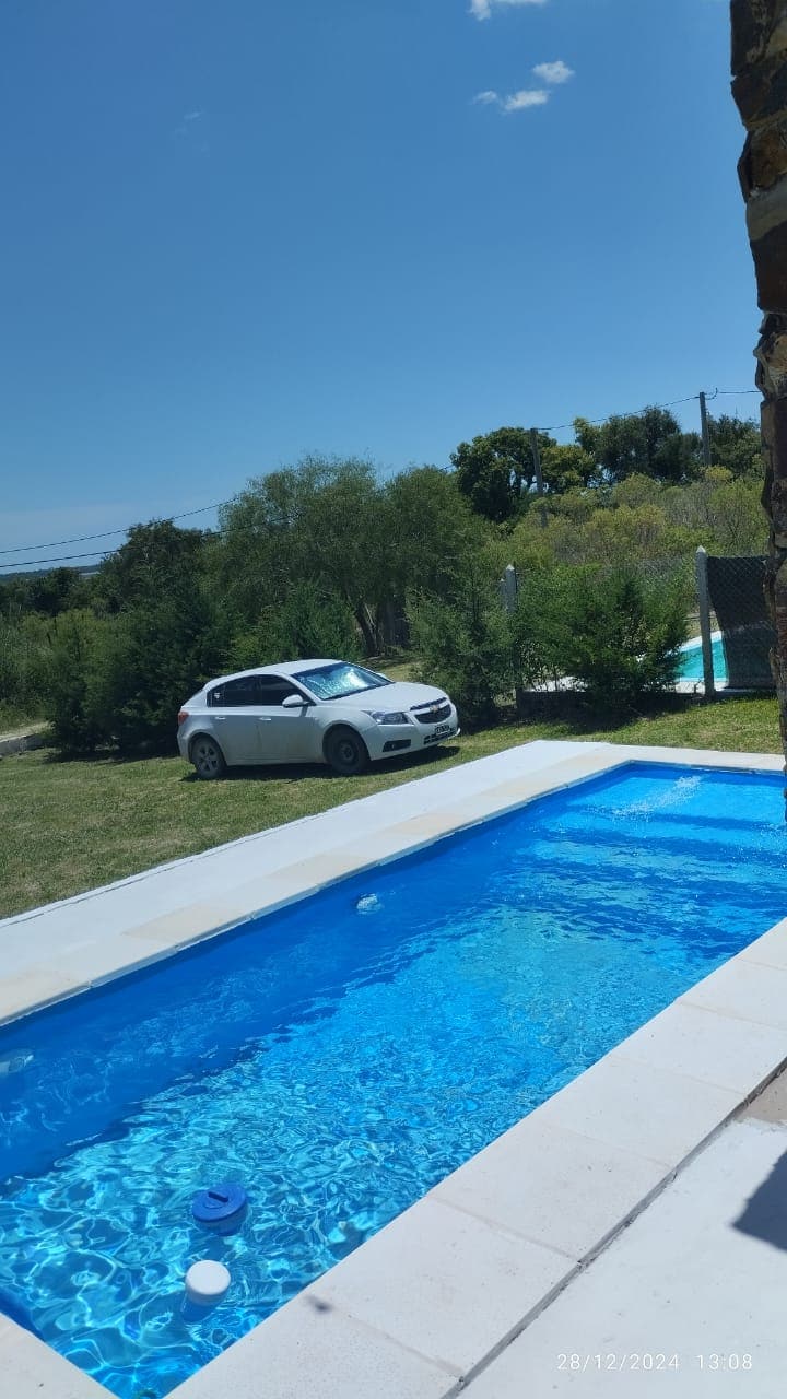 A well-maintained swimming pool is visible, featuring clear blue water and surrounded by a grassy area. A vehicle is parked nearby, and lush greenery is present in the background, adding to the tranquil outdoor setting.