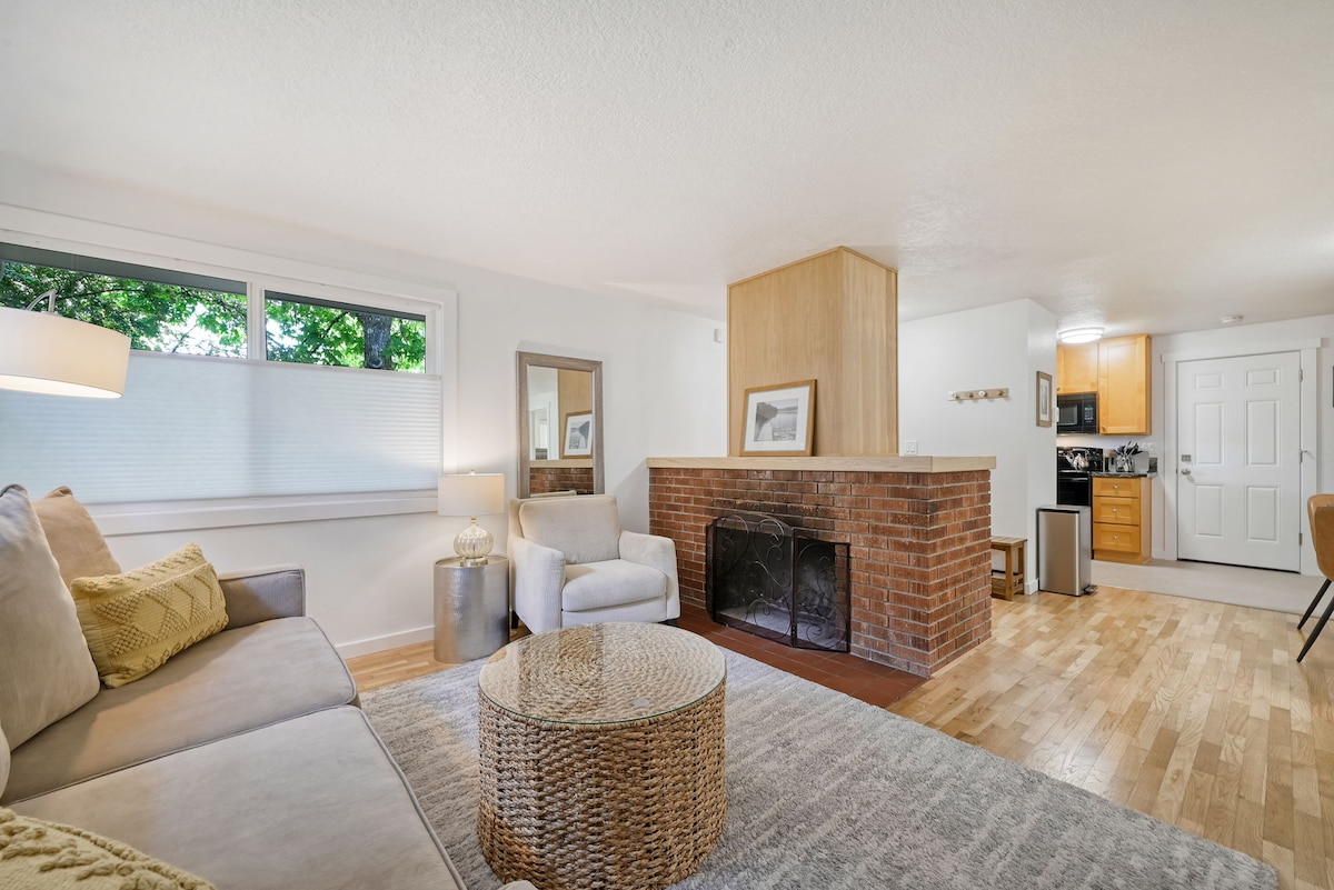 A cozy living area features a light-colored sofa and an accent chair beside a round coffee table. The brick fireplace serves as a focal point, with natural light streaming through large windows. A seamless view of the kitchen is visible in the background.