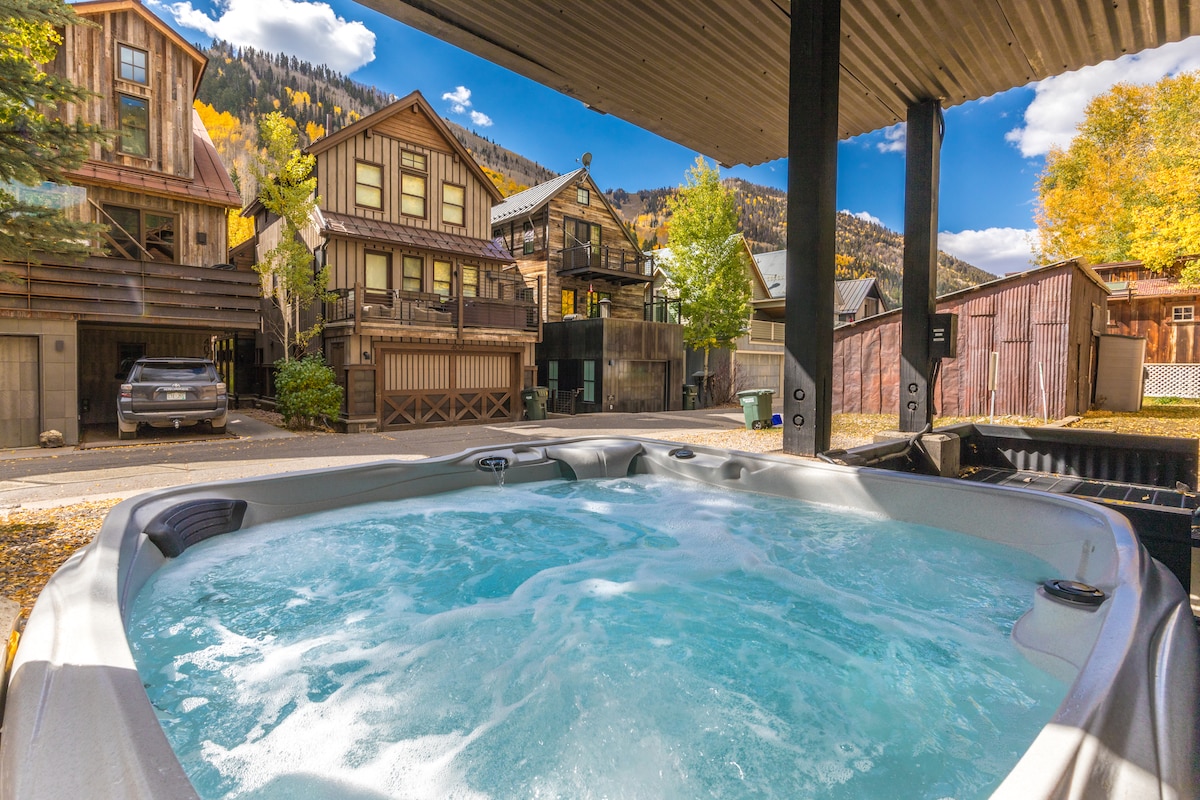 A bubbling hot tub is positioned under a covered patio, inviting relaxation. Surrounding structures include charming mountain homes, with golden autumn foliage visible in the background. Clear blue skies provide a bright contrast to the scene.