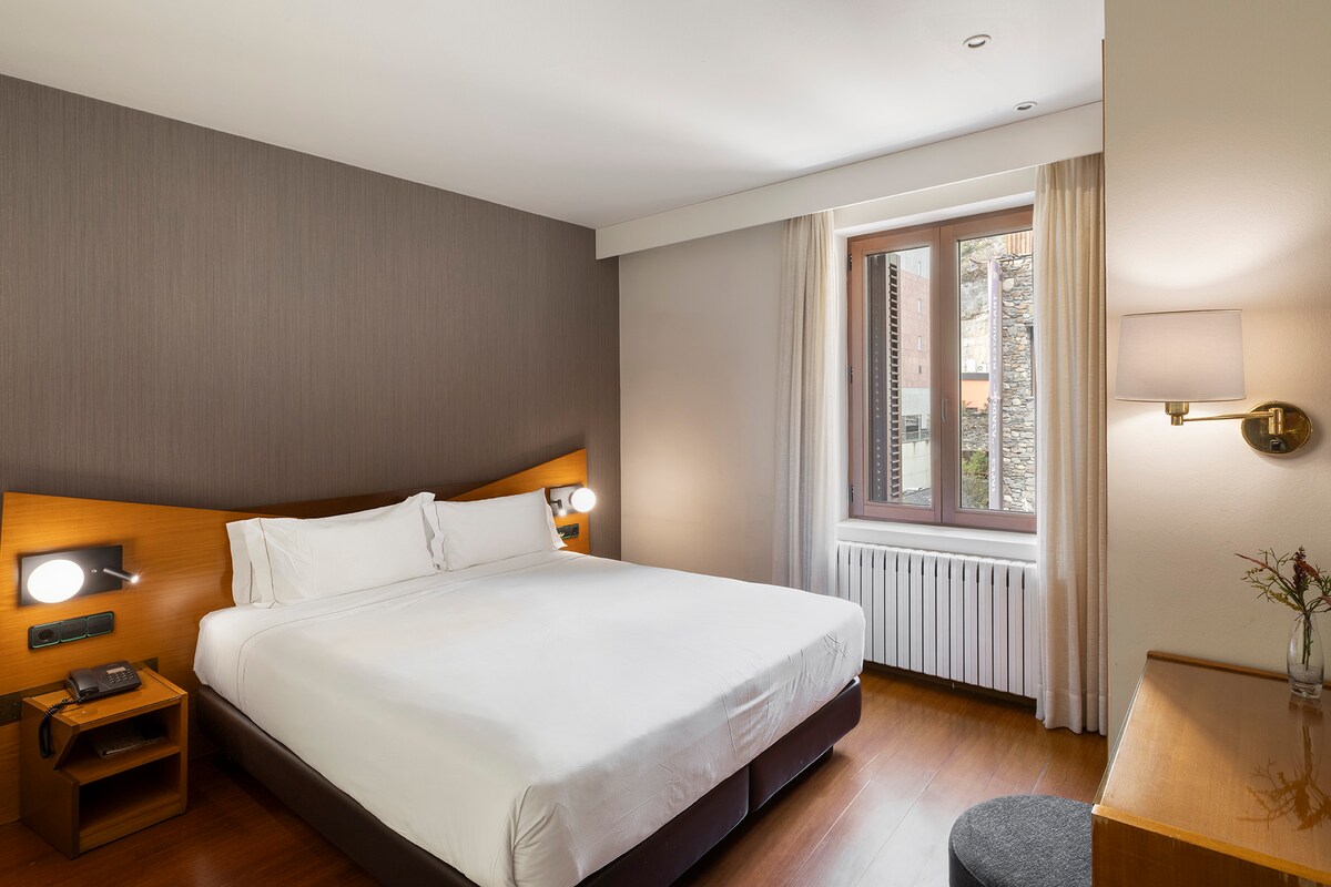 A bright room features a spacious bed with crisp white linens and matching pillows. Natural light enters through a window dressed with light curtains. A wooden desk is placed against the wall, accompanied by a round, upholstered chair. Wall-mounted lights and a bedside table enhance functionality.