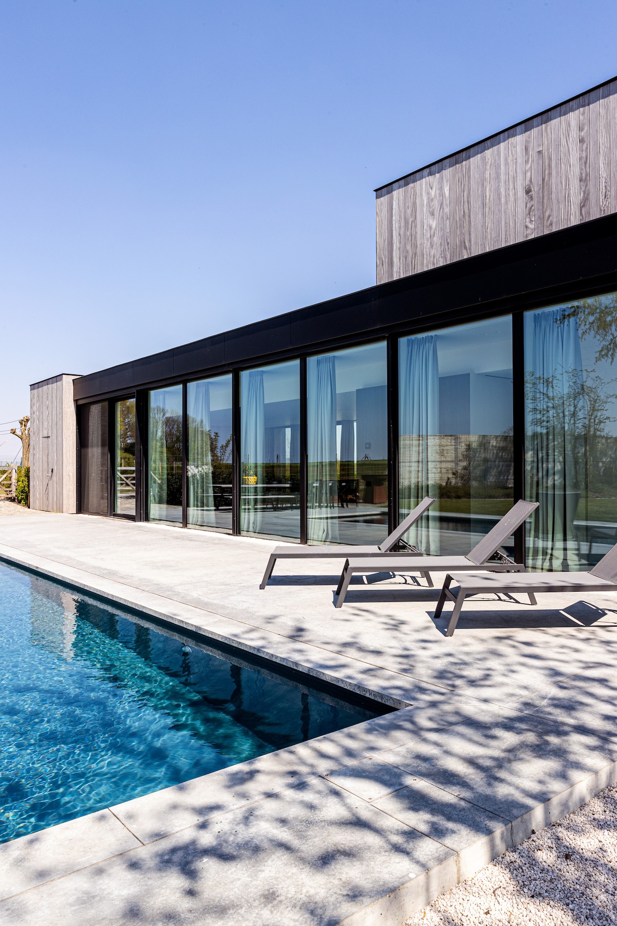 A modern home exterior is visible, featuring large glass doors that provide a view of the outdoor area. Lounge chairs are positioned along the edge of the heated swimming pool, which reflects the clear blue sky. Natural light enhances the spacious feel of the setting.