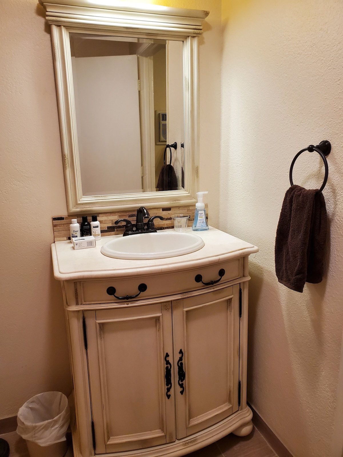 A bathroom space features a cream-colored vanity with intricate detailing and a round sink. Above the sink, a framed mirror reflects the softly lit area. An elegant towel hangs from a metal ring, and personal care items are neatly arranged on the countertop.
