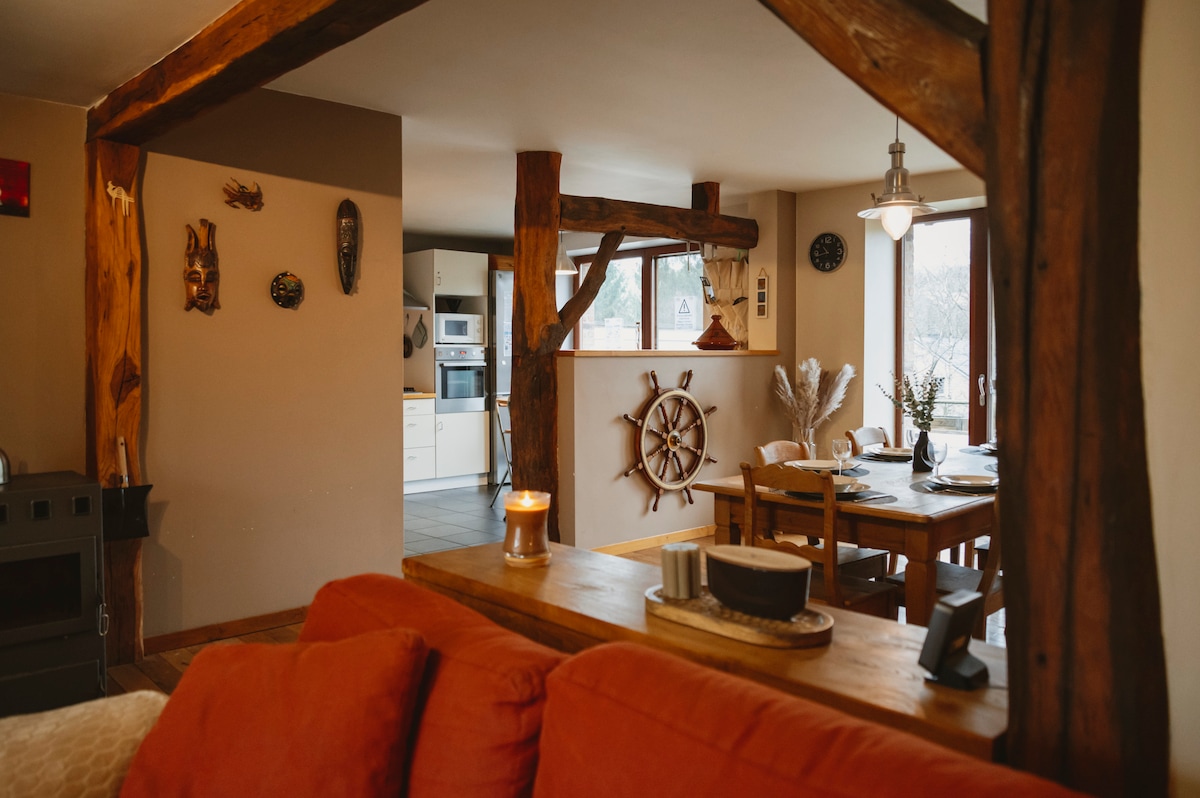 A rustic yet modern living space with exposed wooden beams frames the cozy area. A comfortable couch is positioned near a low wooden table, while a dining table with chairs is visible in the background. Natural light streams through the windows, enhancing the inviting ambiance.