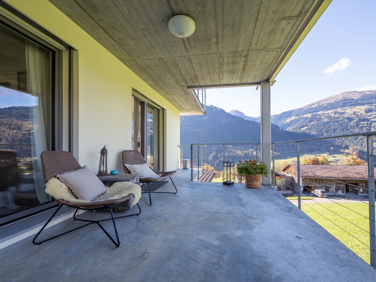 A spacious terrace is furnished with two comfortable chairs and features a view of surrounding mountains and valleys. The concrete flooring adds a modern touch, while a potted plant provides a splash of greenery. Large sliding doors lead into the living area.