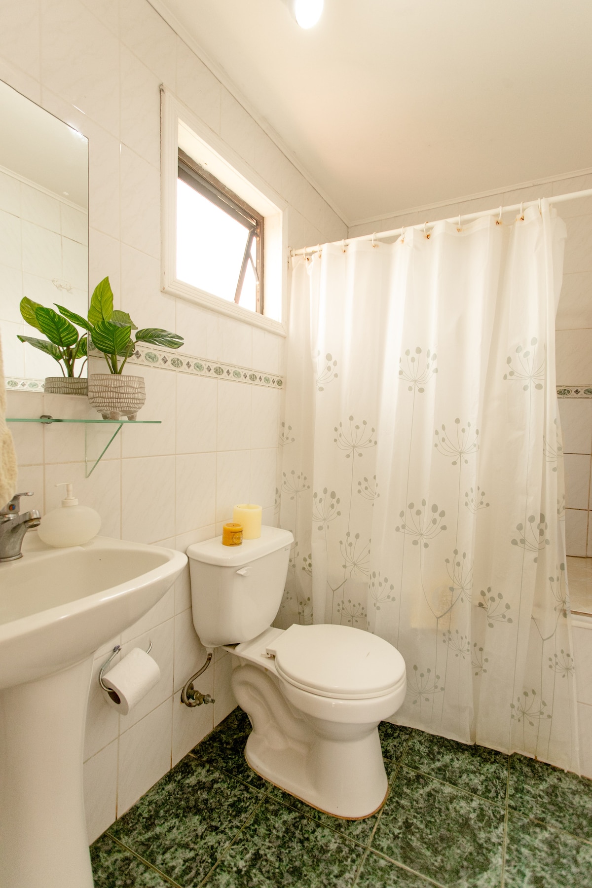 A clean bathroom features white tiled walls and green patterned flooring. A shower area is enclosed by a light curtain adorned with subtle floral designs. A mirror hangs above a sink with a small plant and a candle placed on a glass shelf.