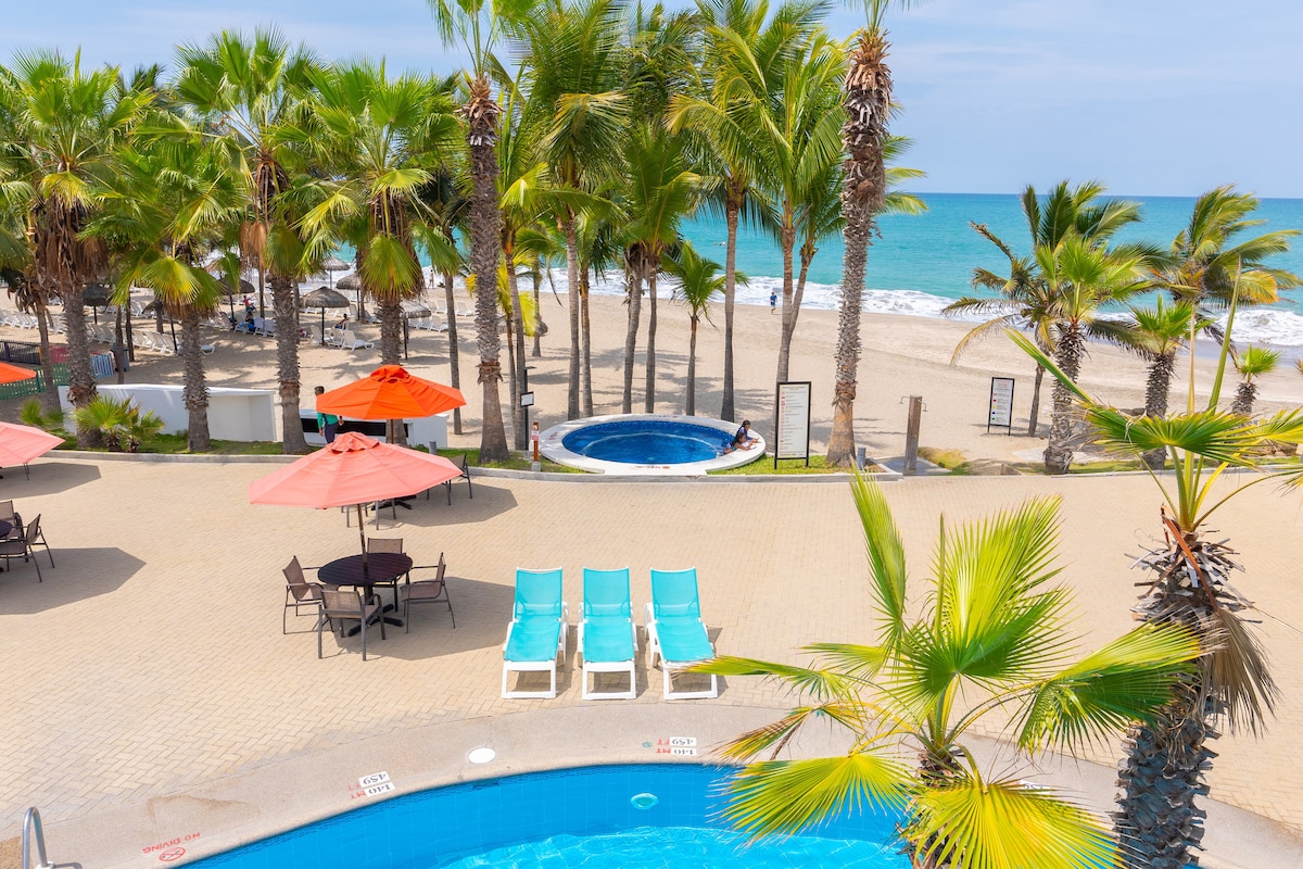 A serene outdoor area features several palm trees surrounding a large pool and a jacuzzi. Beach chairs in light blue are arranged neatly beside the pool, while orange umbrellas provide shade. The beach and ocean are visible in the background, enhancing the tranquil setting.