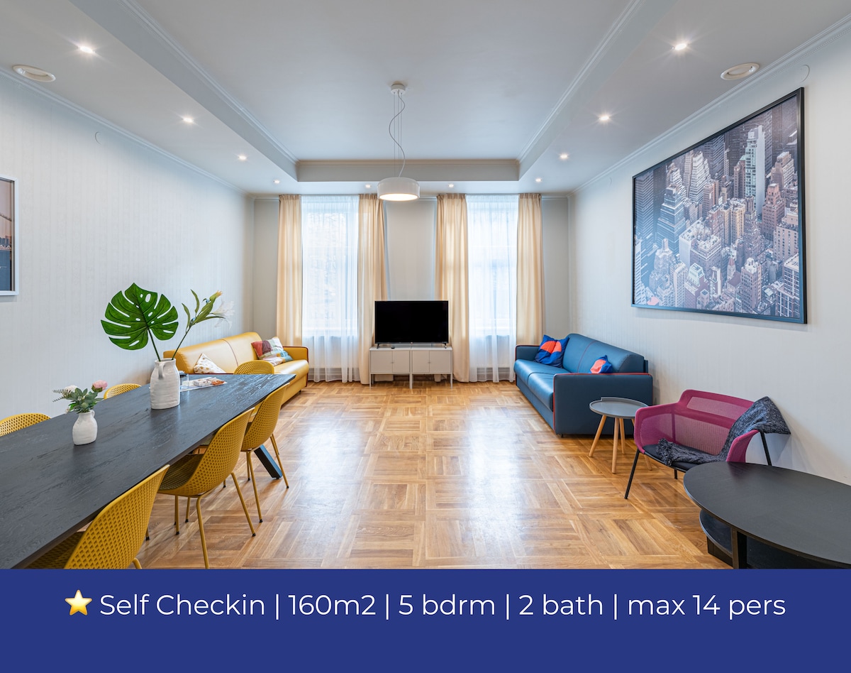 The living room features a spacious layout with a long dining table and colorful chairs, complemented by two sofas. A large TV is positioned against a wall, while natural light filters through tall windows dressed in light curtains, enhancing the room's welcoming feel.
