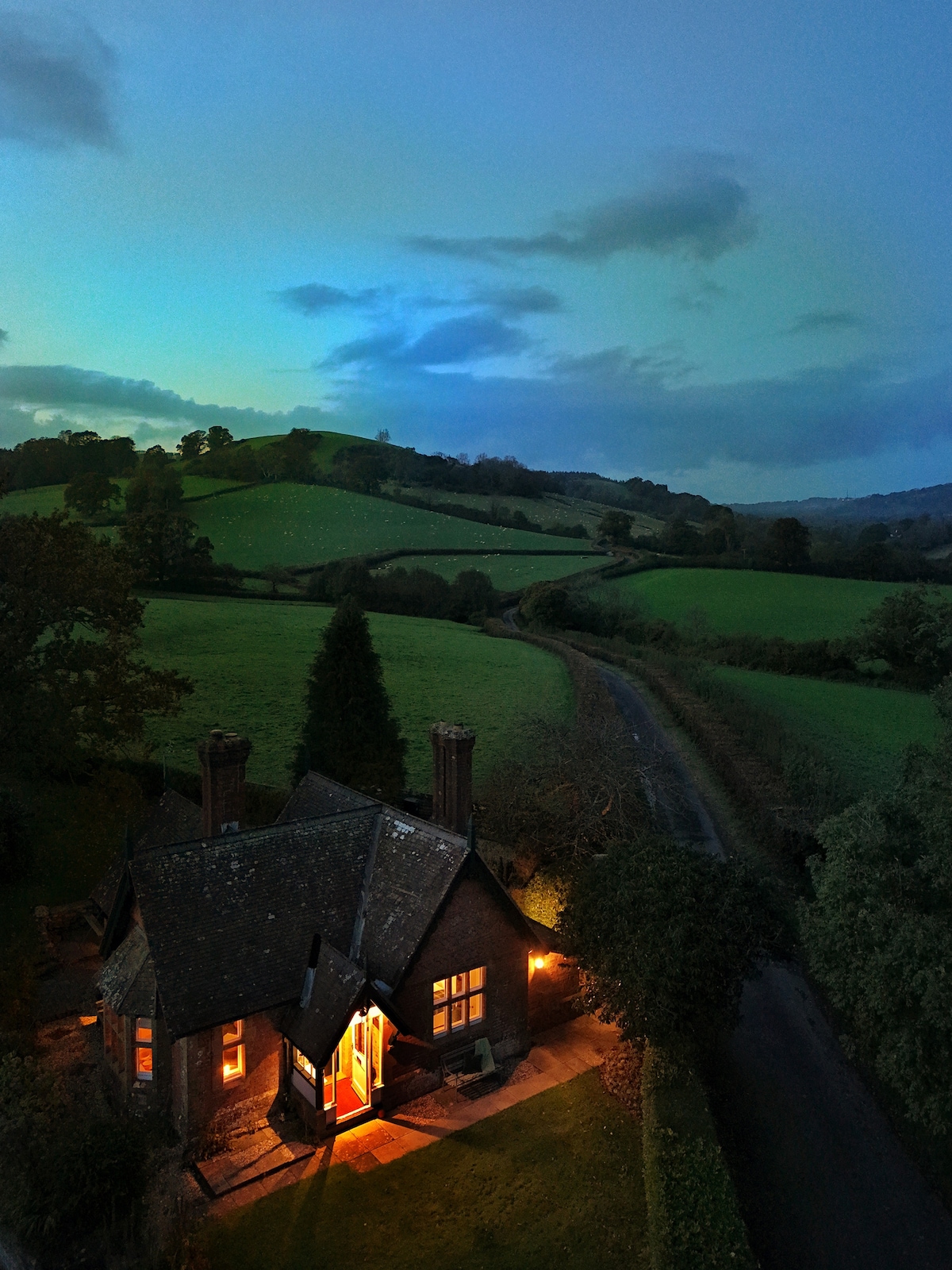 An aerial view captures East Lodge at twilight, nestled among rolling green hills. Warm light emanates from the cozy cottage exterior, contrasting against the evening sky. A winding lane leads to the property, inviting exploration of the tranquil countryside.
