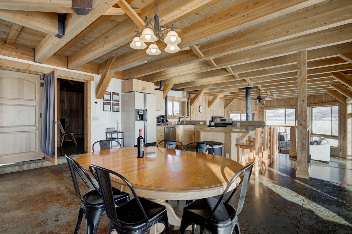 An inviting dining area features a large round wooden table surrounded by black metal chairs. A spacious kitchen with modern appliances is visible in the background, illuminated by natural light streaming through large windows. Rustic wooden beams add character to the open layout.