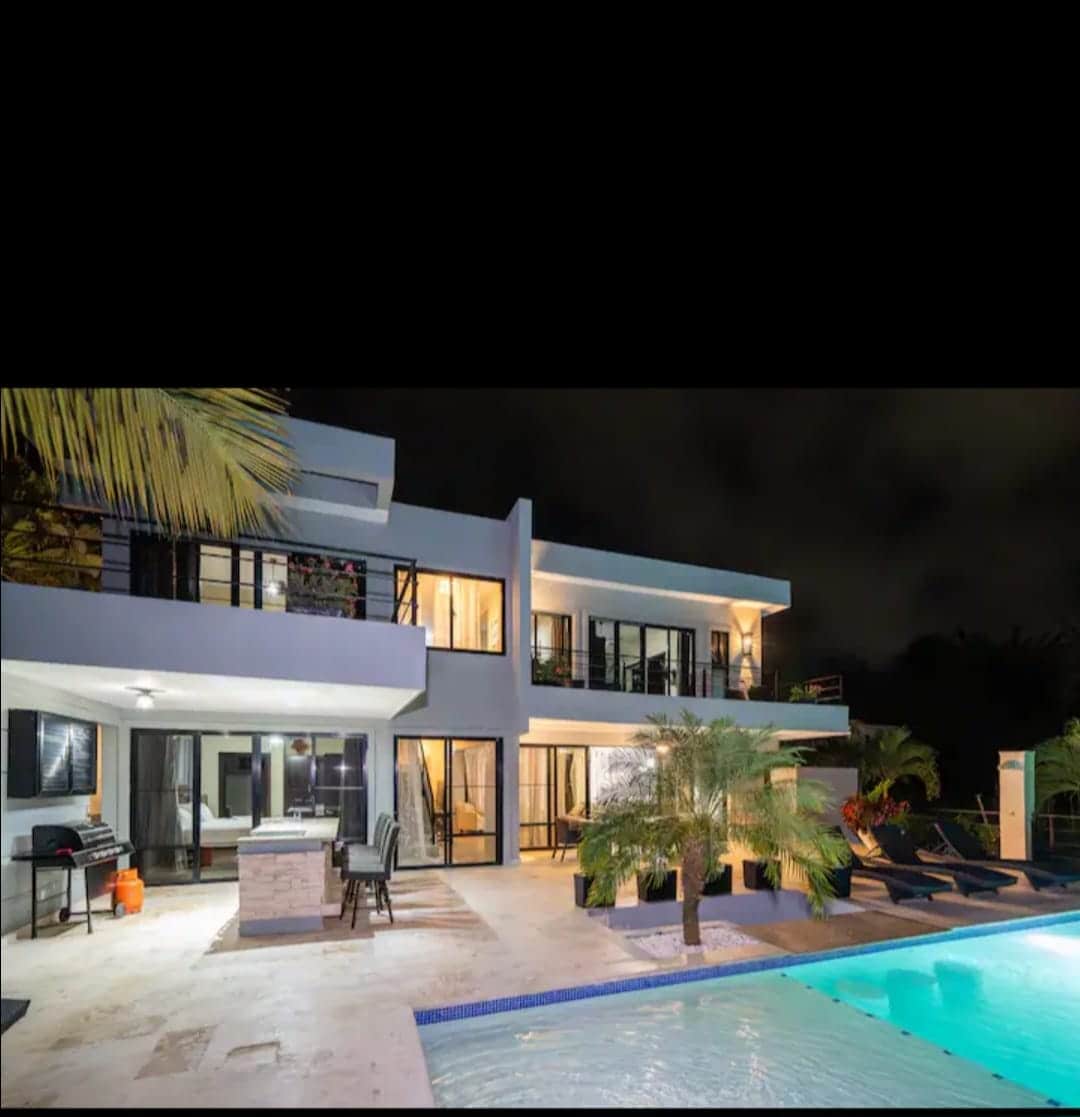 A modern villa exterior is showcased, featuring large glass windows and sliding doors that offer views of the outdoor terrace and swimming pool. Lush palm trees are positioned near the pool, contributing to the tropical ambiance, while outdoor seating is visible on the patio.