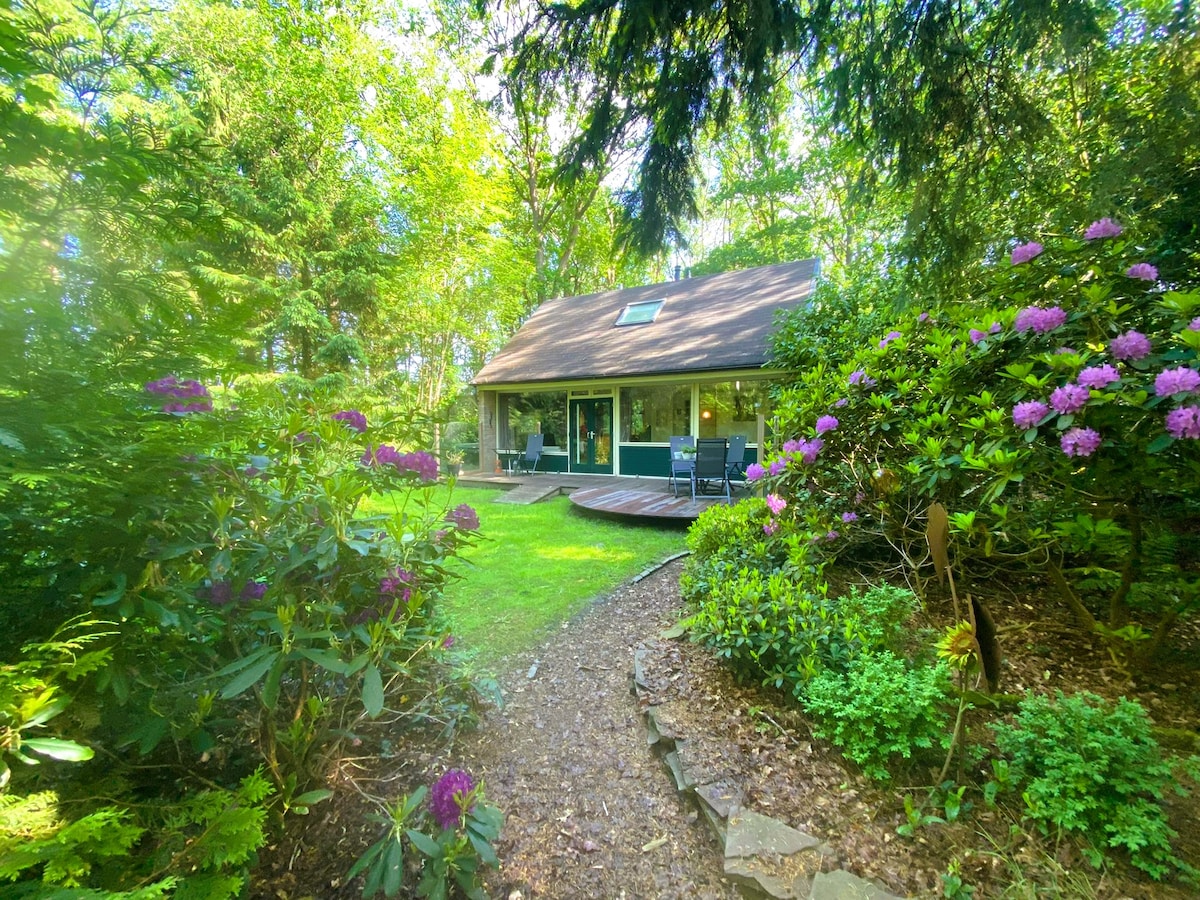 A charming house surrounded by lush greenery and colorful flowers is visible. A spacious deck extends from the front, providing a welcoming entryway. The natural landscape is enhanced by tall trees, creating a serene and private atmosphere, perfect for relaxation in nature.