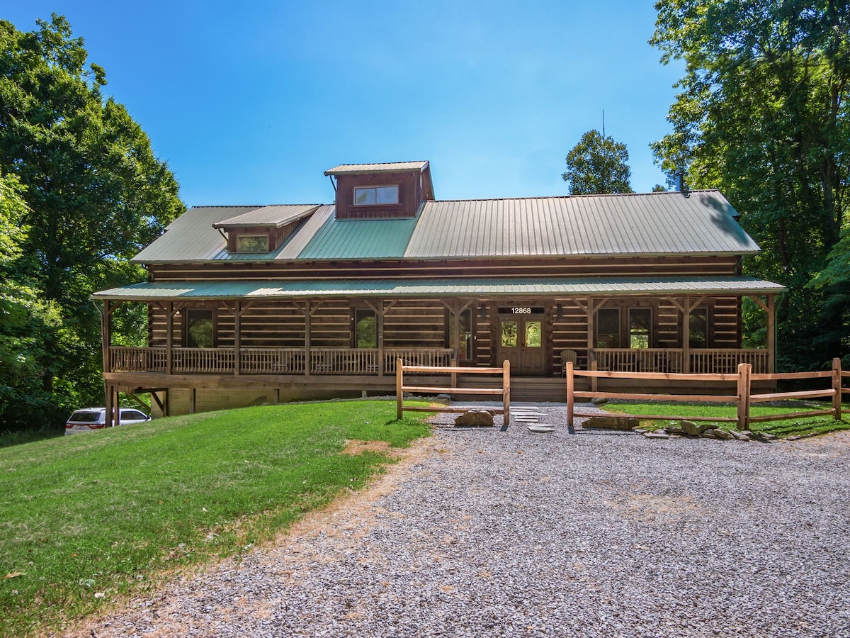 A log cabin featuring a sloped metal roof and large front porch extends across the facade. The structure is framed by lush greenery and a manicured lawn. A gravel pathway leads to the entrance, where wooden fencing adds rustic charm.
