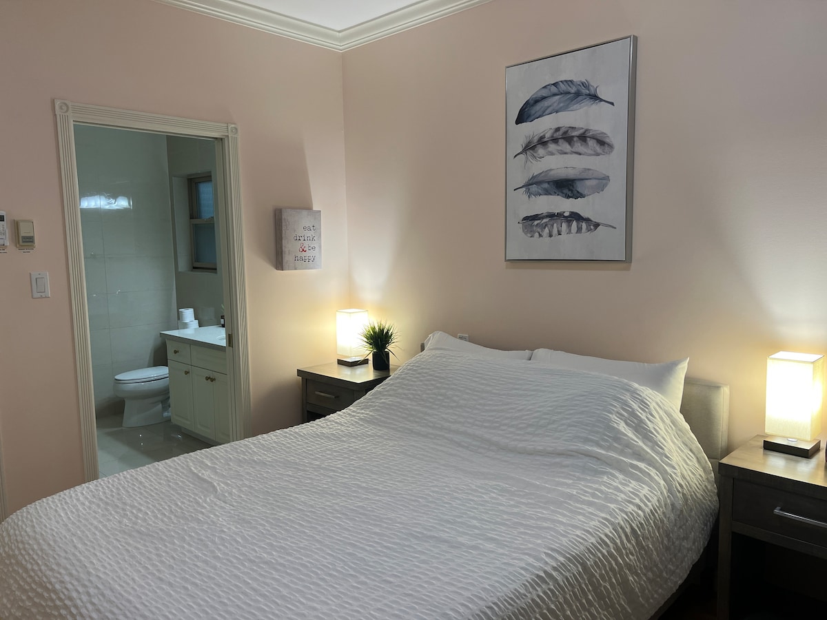 A private bedroom features a spacious bed adorned with a textured white coverlet. Nightstands with lamps are situated on either side. A framed artwork of feathers hangs above the bed, and an ensuite bathroom is visible in the background through an open door.