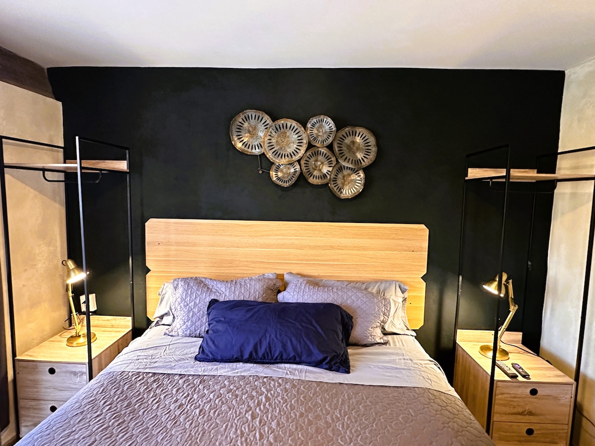 A cozy sleeping area features a spacious bed with a neutral bedspread and a dark blue accent pillow. Two modern nightstands with lamps flank the bed. Decorative wall art, composed of circular metal pieces, adds a stylish touch against the dark backdrop.