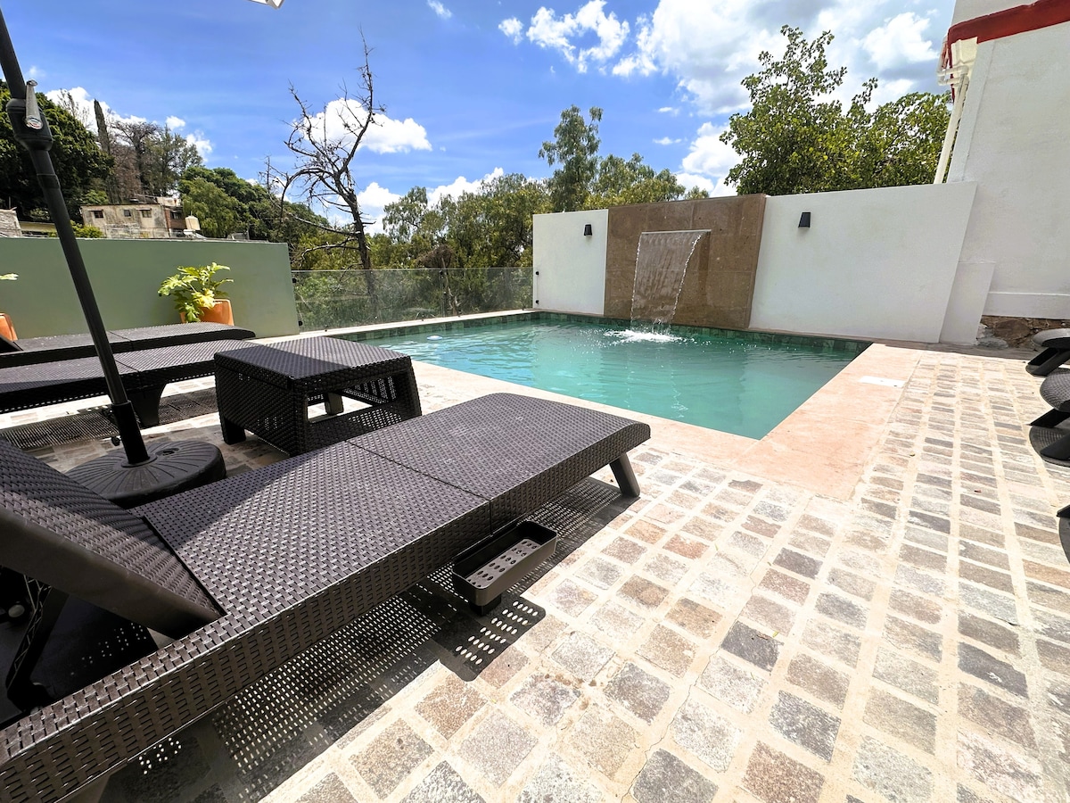 A modern outdoor space is highlighted by a rectangular pool featuring a gentle waterfall. Lounge chairs are arranged along a stone-paved patio, complemented by greenery and views of surrounding trees. Bright skies reflect off the water, creating a serene atmosphere conducive to relaxation.