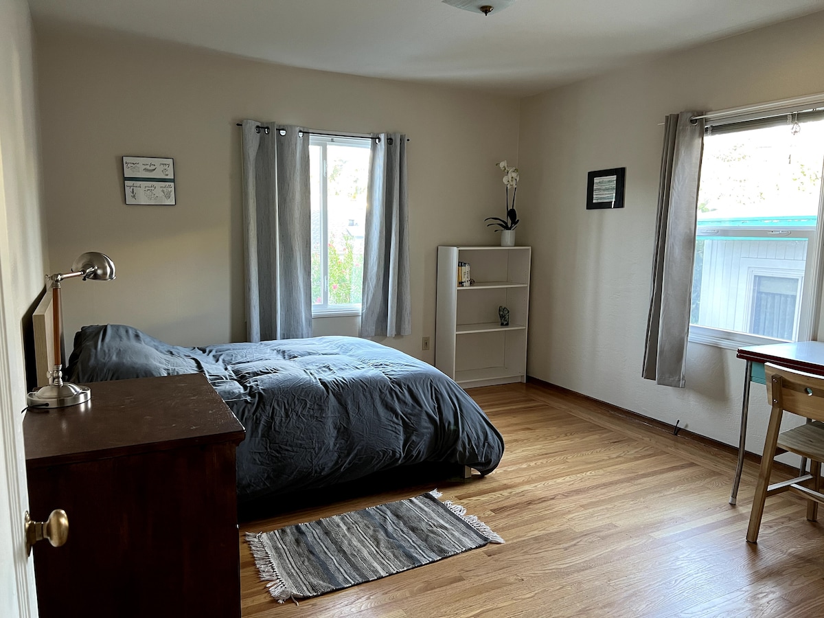A spacious master bedroom is brightened by natural light from two windows, featuring a large bed with a gray comforter. A wooden dresser and shelf provide storage, while a simple desk and chair create a workspace. The hardwood floor adds warmth to the room.