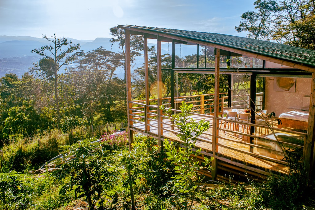 A wooden structure is depicted with large glass walls, surrounded by lush greenery and a scenic view of the mountains in the background. The sunlight shines on the deck, which features wooden railings and slight elevation above the surrounding landscape.