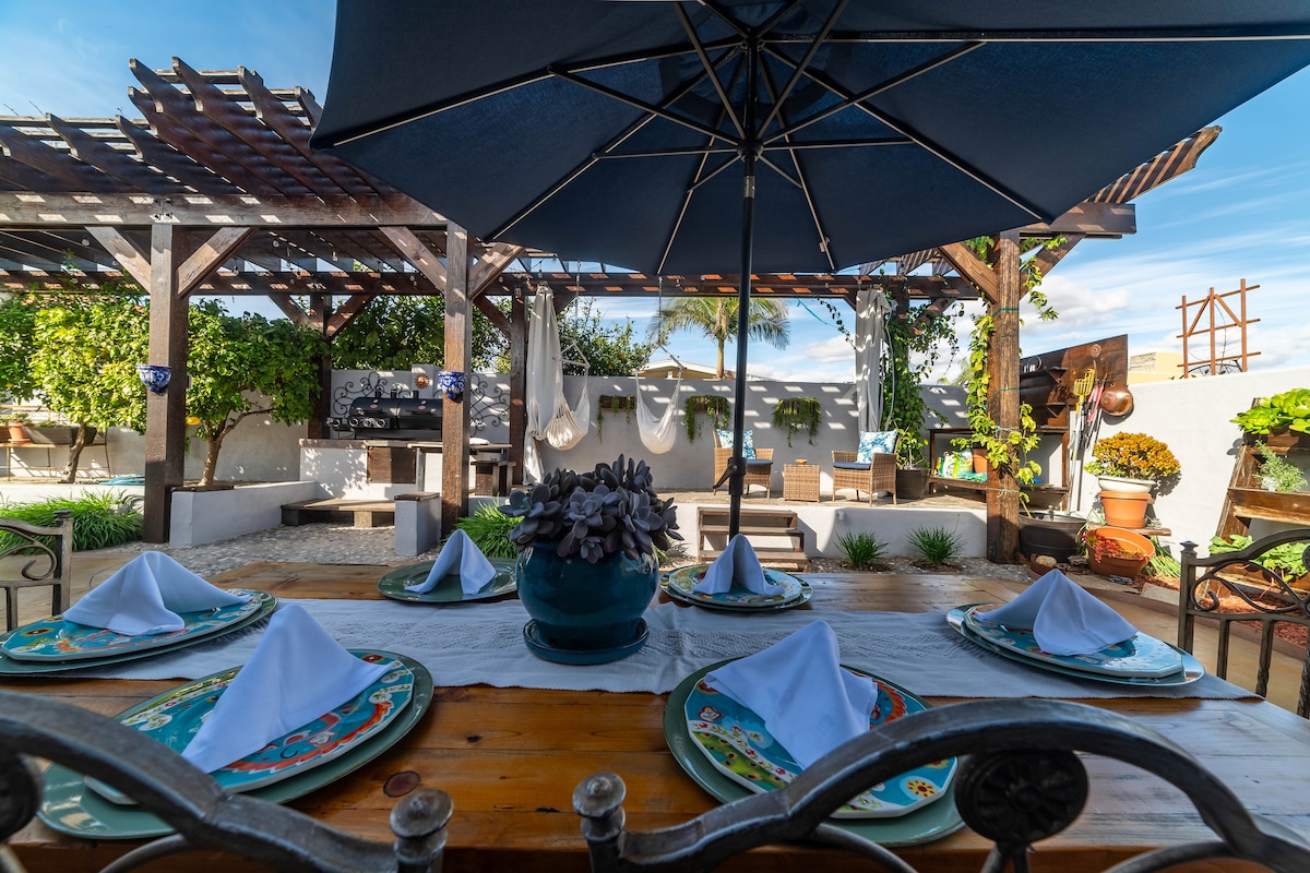 An outdoor dining area features a large wooden table set with decorative dishes and neatly folded napkins. An umbrella provides shade above the table. Surrounding greenery and hanging hammocks create a tranquil atmosphere, while a pergola frames the space, enhancing the inviting outdoor setting.