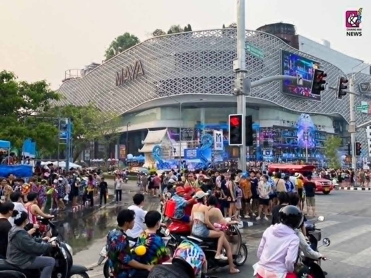 A busy street scene captures the Maya Shopping Mall, characterized by its unique architectural design. Numerous pedestrians and motorbikes are visible, with colorful crowds gathered around a festive event, showcasing the vibrant atmosphere of the area. Traffic signals and urban greenery frame the bustling setting.