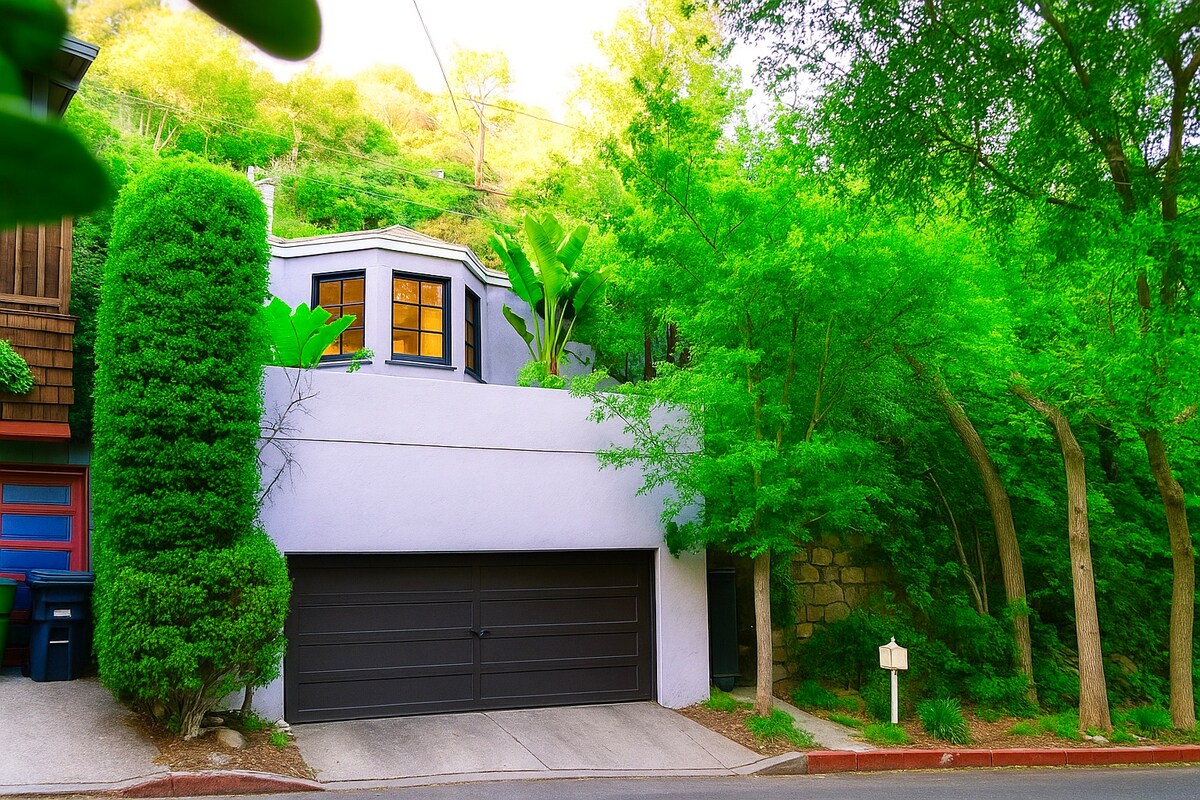 The exterior of a hillside home is visible, featuring a light-colored façade and a spacious garage below. Lush greenery surrounds the property, with vibrant trees and plants enhancing the natural setting, creating a serene atmosphere in the Hollywood Hills.