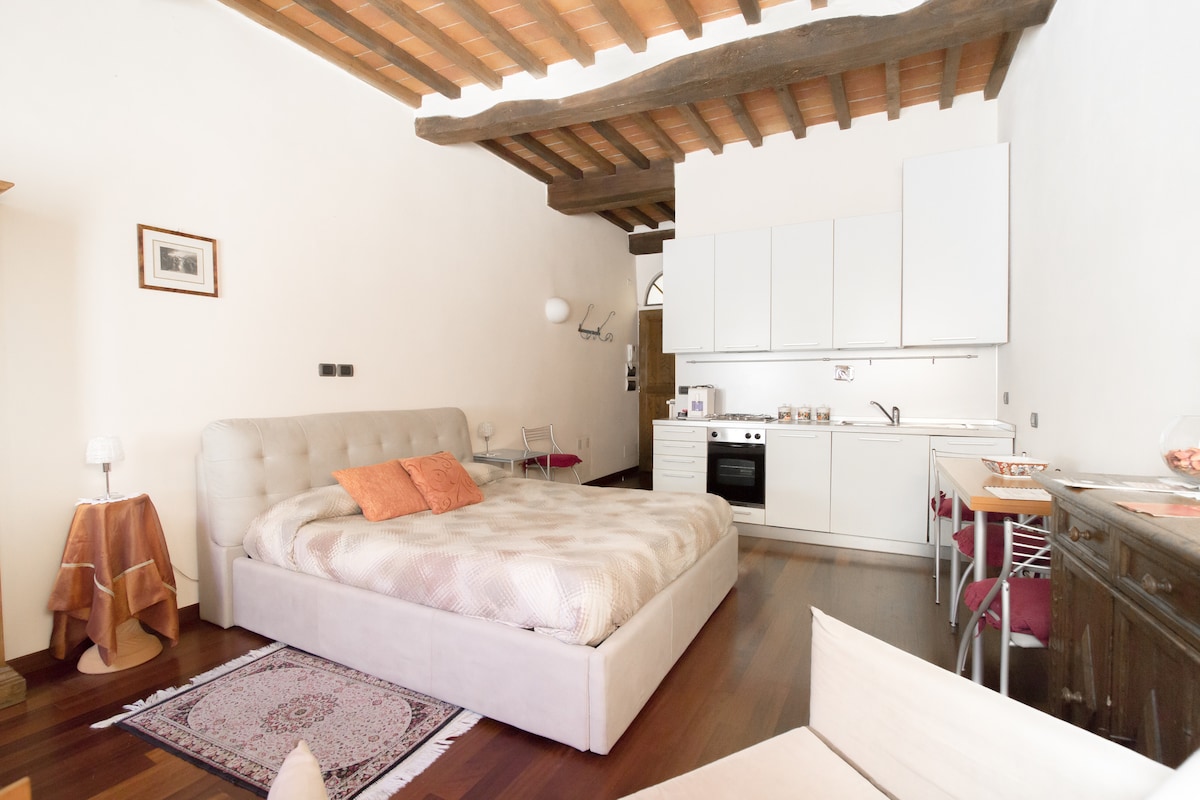 The junior suite features a spacious bed paired with soft pillows, adjacent to an open kitchenette equipped with modern appliances. Light wooden beams frame the ceiling, and a dining area with minimalist chairs is visible. A subtle area rug adds warmth to the hardwood floor.