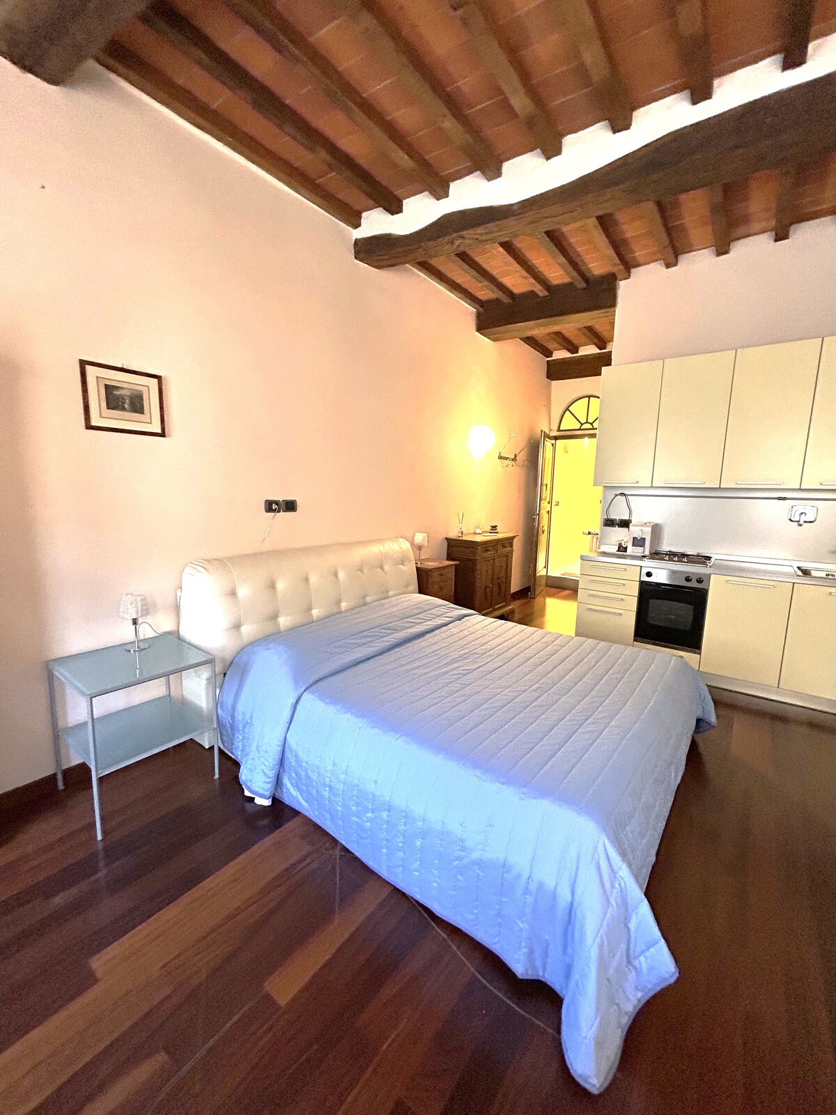 A junior suite features a spacious bed covered with a light blue quilt. The rustic wooden ceiling enhances the space, which includes a well-equipped kitchen area and a small table beside the bed. Soft lighting contributes to a warm ambiance.