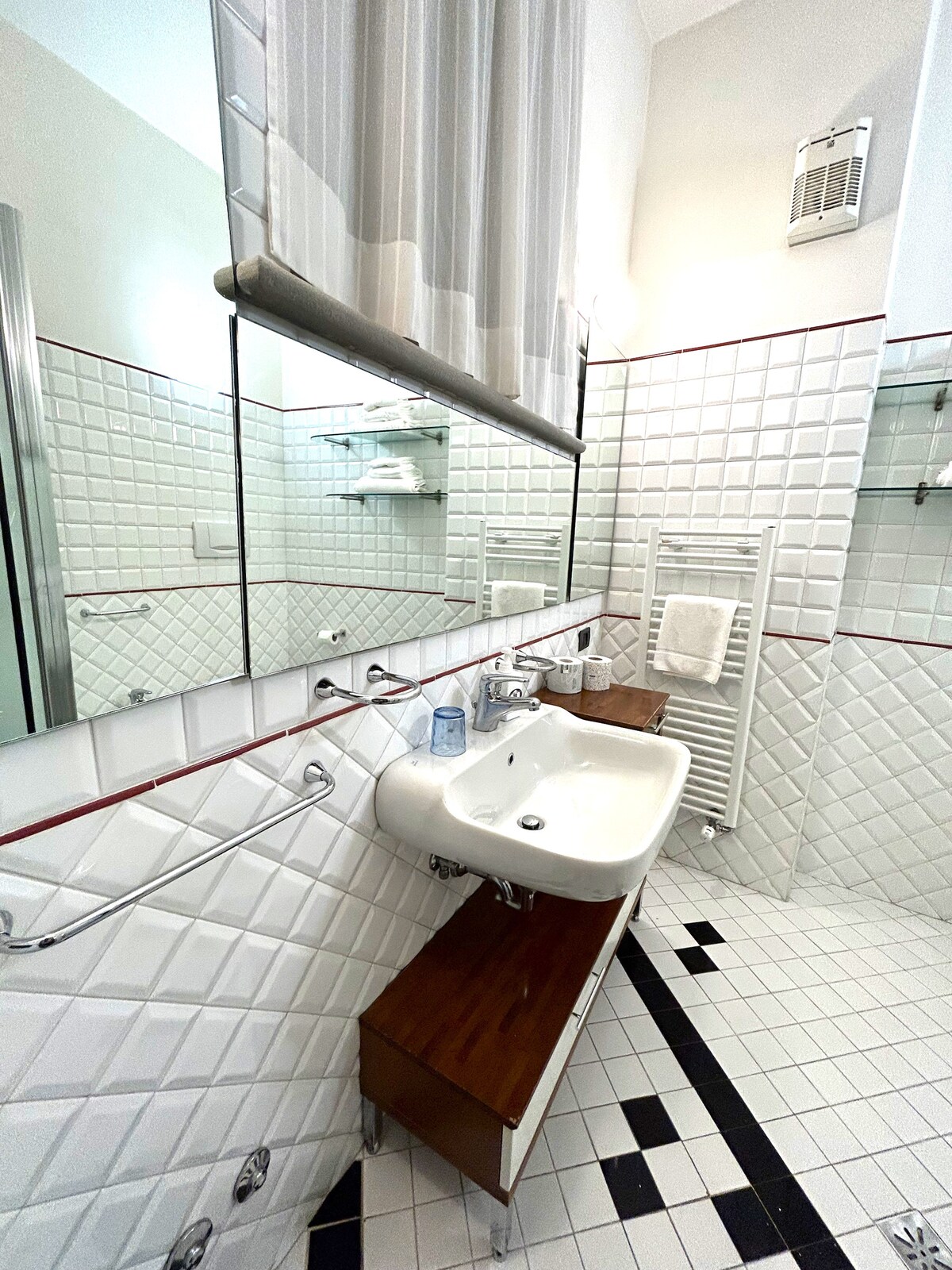 The bathroom features white tiled walls with decorative accents and a spacious layout. A large mirror reflects the area, which includes a modern sink and wooden vanity. Fresh towels are neatly arranged, and the flooring showcases a contrasting black and white tile pattern.