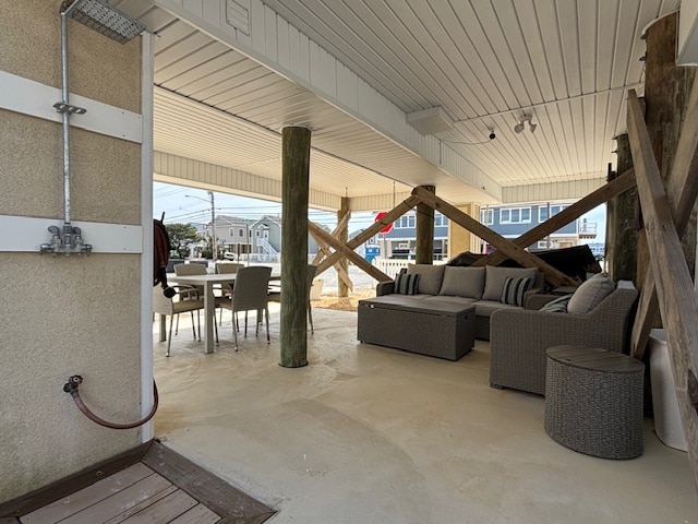 An outdoor lounge area is presented under the house, featuring a comfortable sectional sofa and a round side table. A dining table with chairs is visible, surrounded by supportive wooden beams. An outdoor rinse station is located to the left, with a beach-like sandy floor.