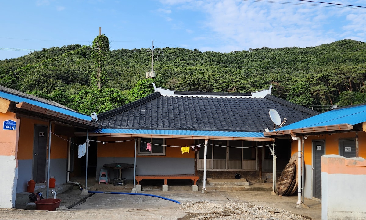 A traditional-style building is set against a lush green hillside. The exterior features a tiled roof and colorful walls. Clothes hang on a line, and a few outdoor items are visible, including a basin and a round table.