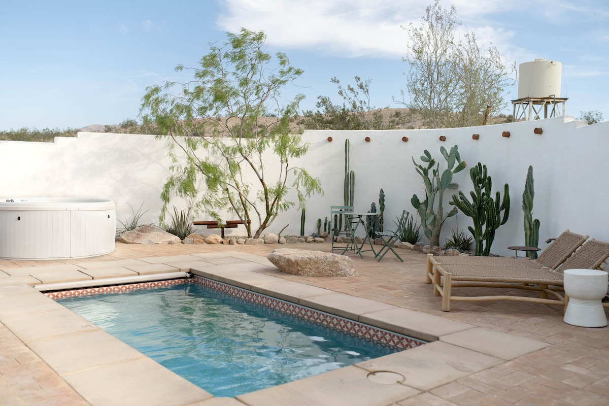 A private courtyard features a heated pool and hot tub surrounded by desert plants, including various cacti. Two lounge chairs are positioned near the pool, while a small table and chairs provide an outdoor seating area. A wall ensures privacy and offers a serene desert backdrop.