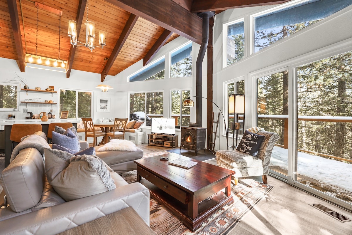The spacious living area features vaulted wooden ceilings and large windows overlooking a serene forest. A comfortable seating arrangement surrounds a wooden coffee table. A fireplace and television add warmth to the inviting space, which transitions seamlessly into the kitchen area.
