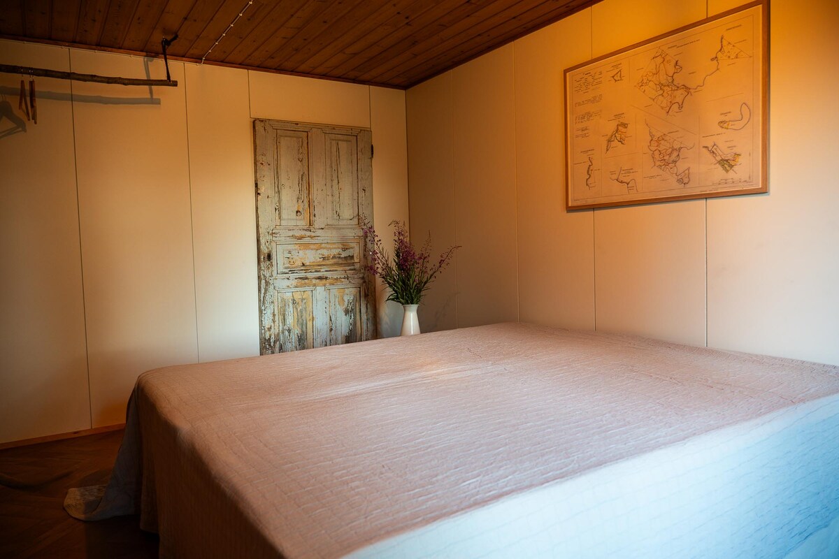 A spacious bedroom is presented with a large bed covered by a light-colored quilt. An old wooden door adds character to the room, while a vase with flowers sits on a simple table, bringing a touch of nature indoors. Soft lighting creates a warm ambiance.