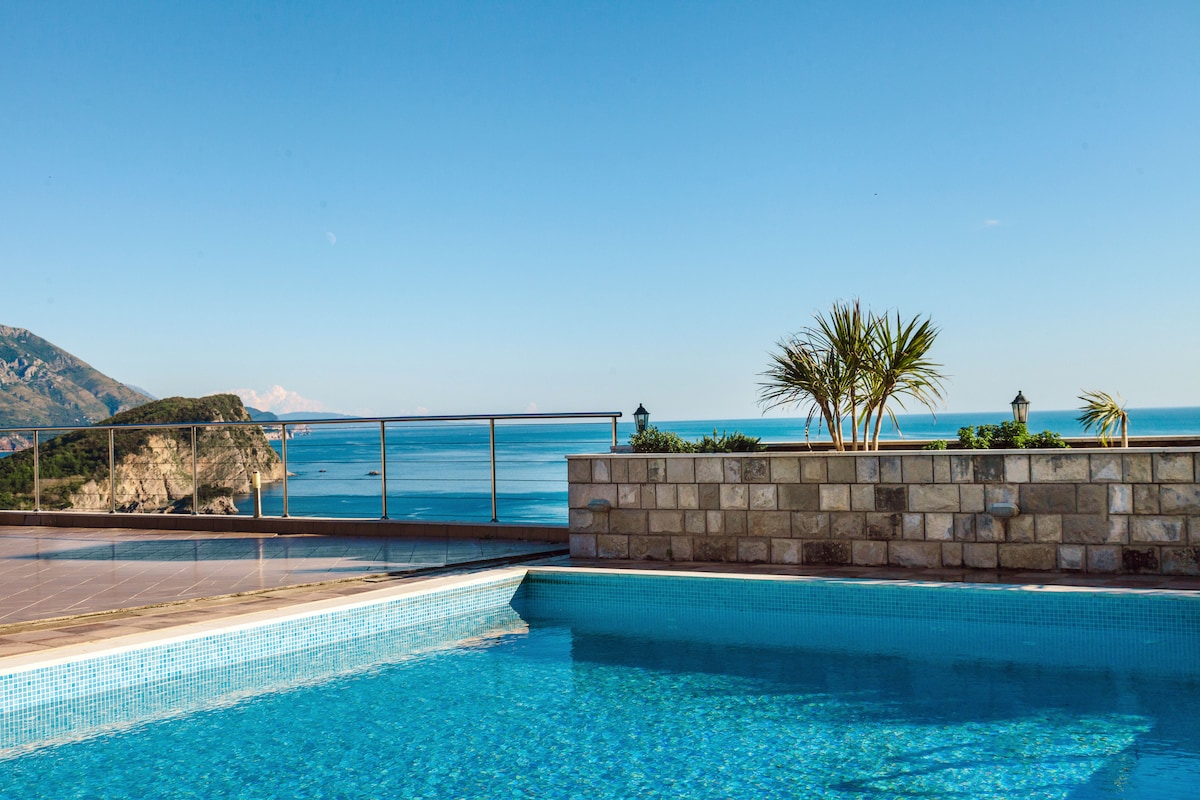 A tranquil swimming pool reflects the clear blue sky and distant mountains, with stunning sea views visible beyond the railing. Palm trees are positioned strategically around the terrace, adding to the serene ambiance.