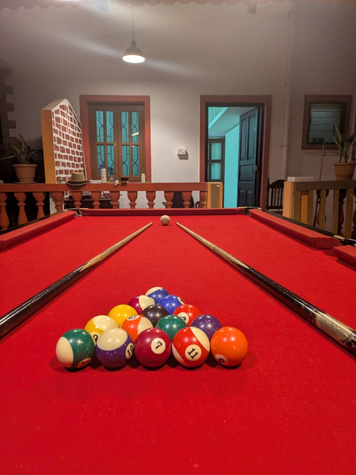 A vibrant pool table is positioned in a spacious room featuring a red felt surface. A collection of colorful billiard balls is arranged in a triangular formation, while two cue sticks rest alongside the table. Doorways and potted plants can be seen in the background.