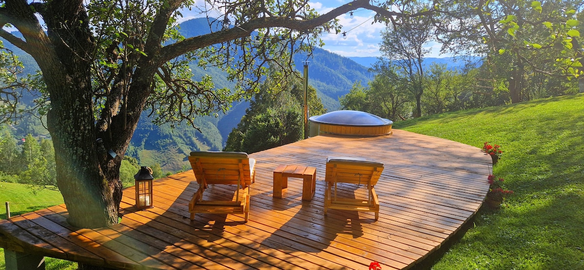 A wooden deck extends into a scenic landscape, featuring two loungers positioned for relaxation. A small table sits between them, while a hot tub is visible in the background. Lush greenery and rolling hills create a serene atmosphere.