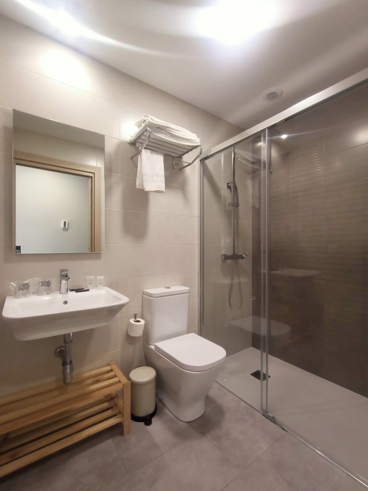 A modern bathroom is featured, showcasing a glass shower enclosure and a sleek sink. A mirror is mounted above the sink, while white towels are neatly arranged on a shelf. A small wooden stool adds a touch of warmth to the space.