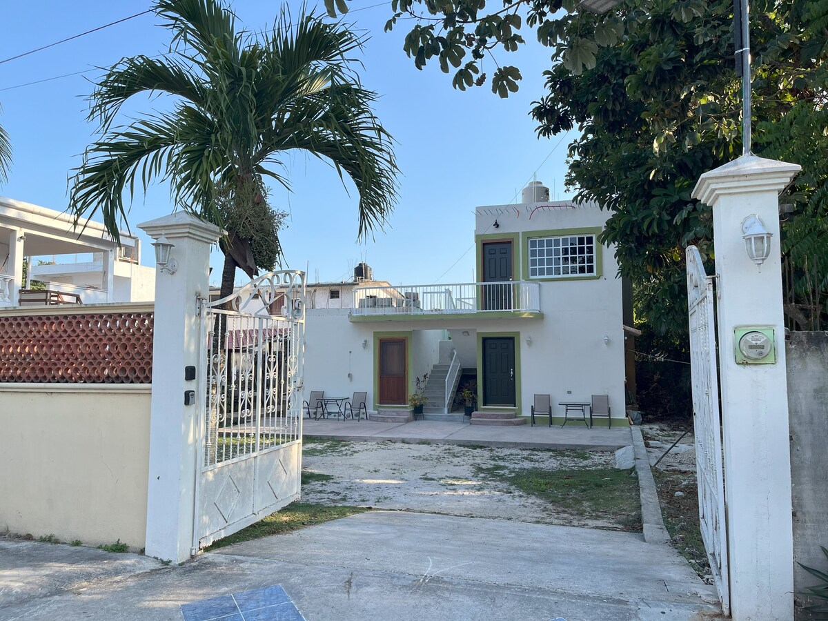 The exterior view of a two-story building is captured, featuring a spacious yard with palm trees. The entrance is marked by a decorative gate, leading to a clear path toward the front door. The structure showcases large windows and a balcony, contributing to its welcoming presence.
