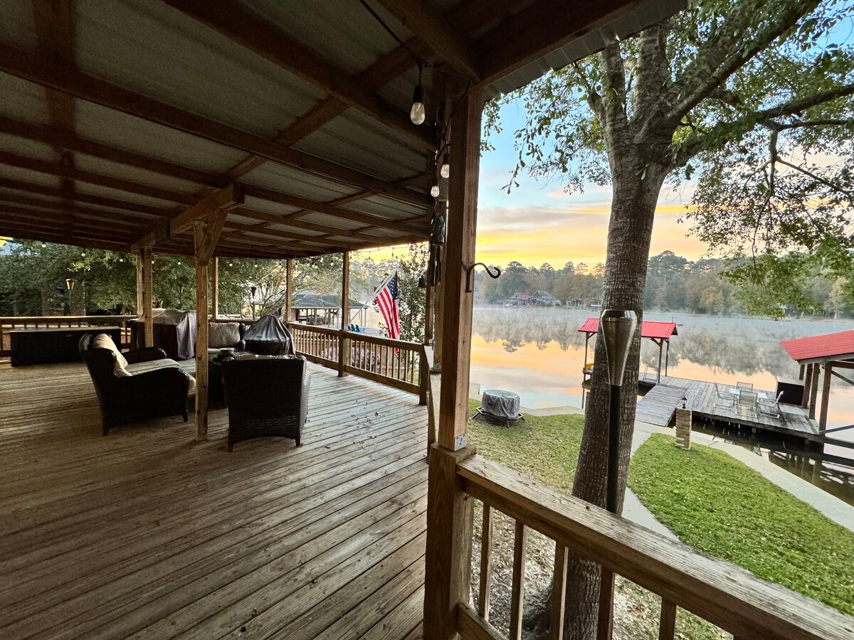 A spacious back porch offers comfortable seating with views of the tranquil lake. Morning light reflects off the water, and a flag gently flutters nearby. The wooden floor and surrounding trees create a cozy, natural atmosphere for relaxing or gatherings.