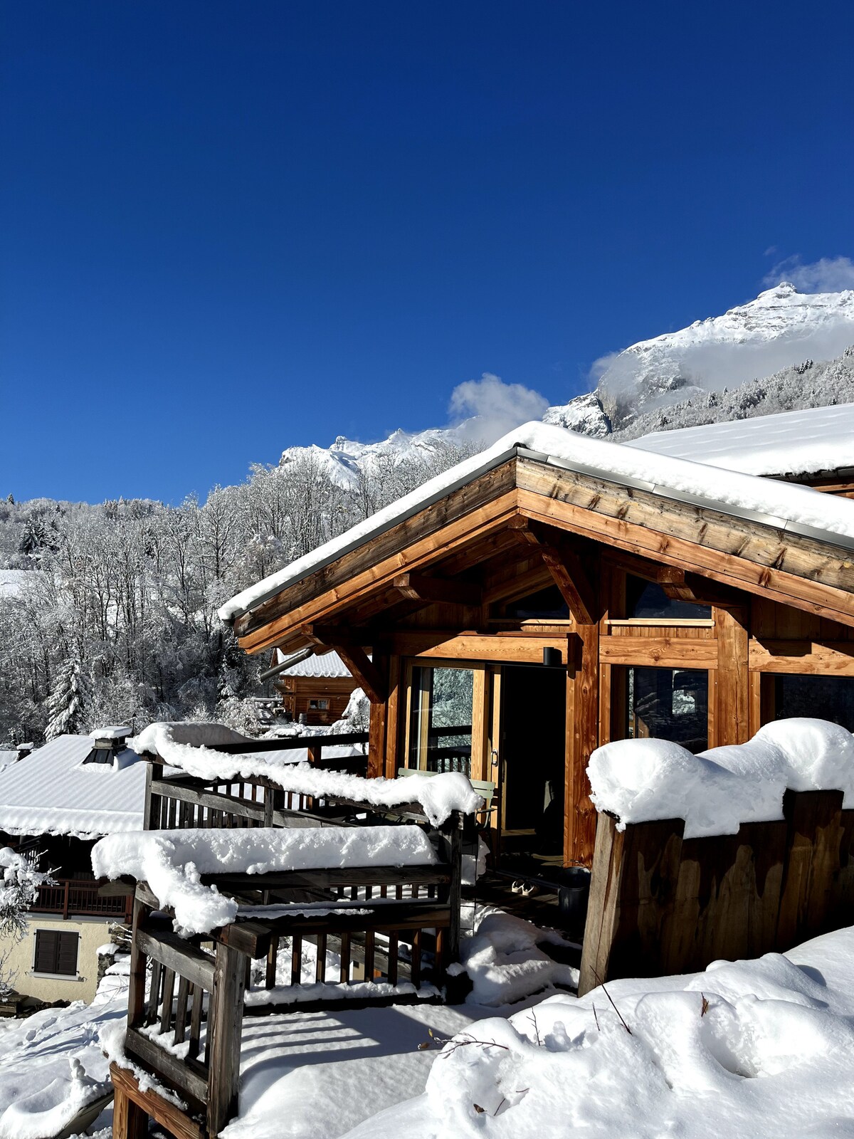 A wooden chalet is nestled among snow-covered rooftops, showcasing a combination of rustic architecture and modern design. Bright sunlight reflects off the freshly fallen snow, while the surrounding mountains are framed by a clear blue sky.