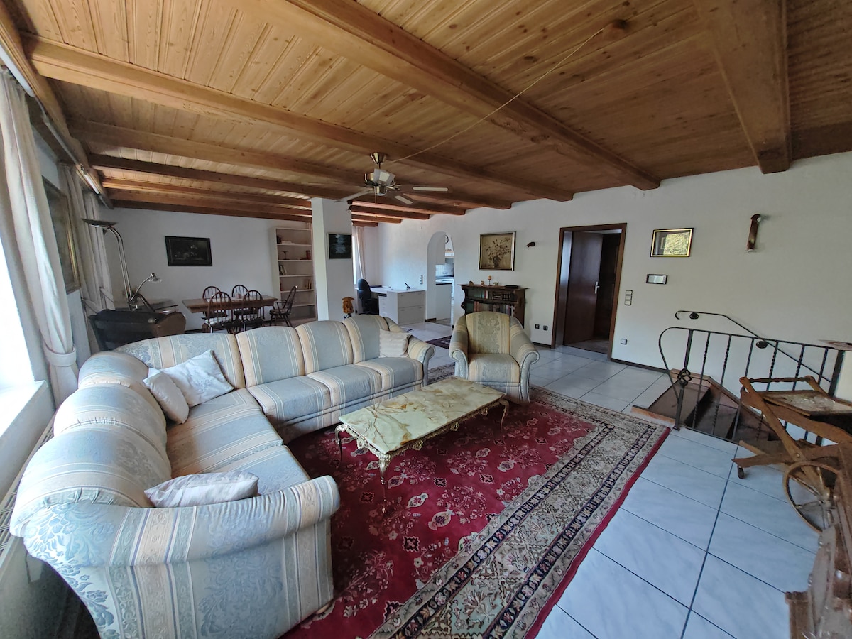 A spacious living area features a large sectional sofa and an antique coffee table atop a decorative area rug. Wooden beams accent the ceiling, while natural light illuminates the room through multiple windows. A dining set is visible in the background, blending with the open layout.