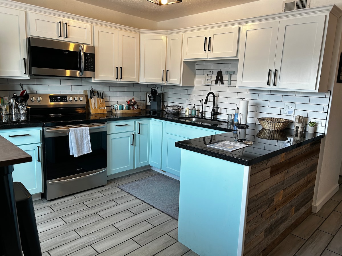 A modern kitchen features white cabinetry complemented by a light blue island and dark granite countertops. Stainless steel appliances are visible, along with a tiled backsplash. Utensils and kitchen tools are neatly arranged, contributing to a functional cooking space.