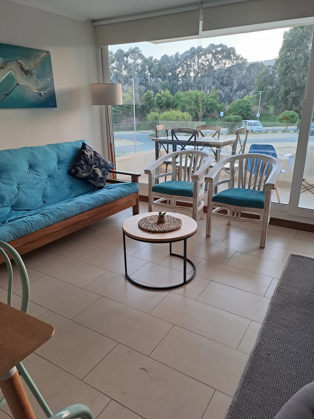 A bright and airy living space is showcased with a turquoise sofa and light wooden chairs surrounding a round coffee table. Natural light fills the room through large glass doors, revealing views of the green landscape outside. A piece of artwork adds a touch of color to the wall.