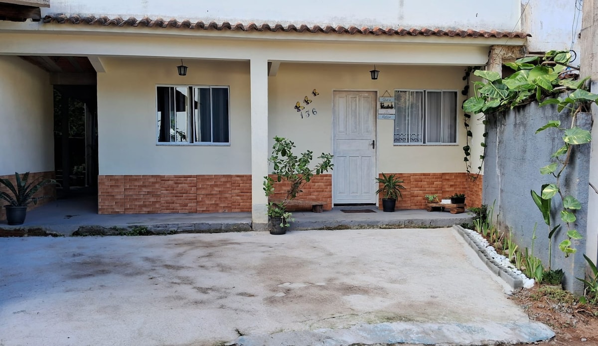 The exterior of a house is displayed, featuring a simple white door and two windows with discreet white curtains. A small potted plant is positioned near the entrance. A concrete area provides space for a car, bordered by a low green hedge on one side.