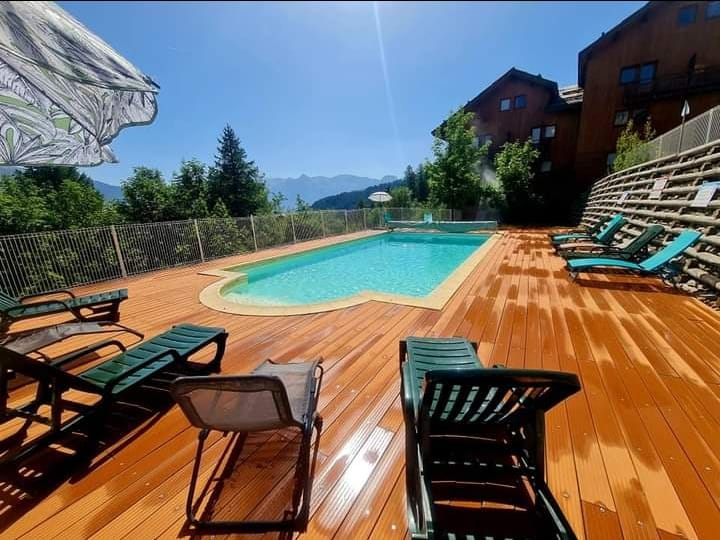 An outdoor pool area is surrounded by wooden decking, featuring several lounge chairs. The pool reflects the clear blue sky and is framed by trees, with mountains visible in the background. Sun umbrellas provide shaded seating options.